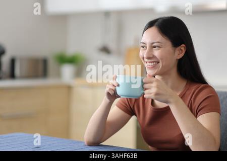 Felice donna asiatica che beve caffè o tè in cucina a casa Foto Stock