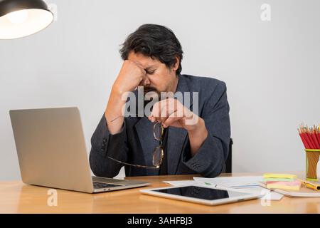 Un uomo indiano stanco che usa il computer portatile per lavorare, un ragazzo in abbigliamento casual sta toccando e massaggiando il ponte del naso, si sente stanco e bruciato, seduto Foto Stock