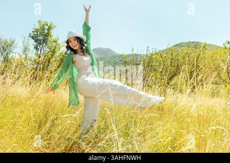 Una giovane donna allegra balla liberamente in un prato dorato circondato da alberi, con montagne sullo sfondo. Concetti di libertà, felicità, natura, Foto Stock