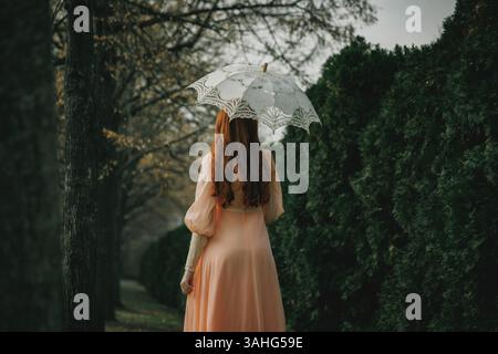 Una donna dai capelli rossi in abito pesca regge un ombrello in pizzo, muovendosi tra alberi e ombre, evocando il fascino del vecchio mondo e il desiderio di tranquillità. Foto Stock