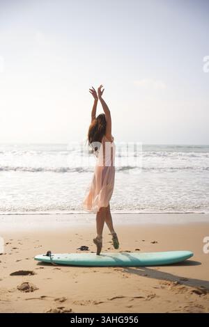 Ballerina spensierata in abito rosa si bilancia sulla tavola da surf in spiaggia tranquilla Foto Stock