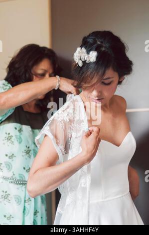 Madre latina che aiuta la sposa figlia il giorno del matrimonio a vestirsi con abiti da sposa con luce soffusa e capelli brillanti. Concetto di novelle e n Foto Stock