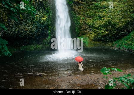 Donna con ombrello rosso in piedi vicino a una lussureggiante cascata in una foresta verde. Silver Falls, Oregon, Stati Uniti Foto Stock