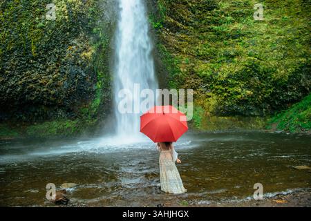 Donna con un ombrello rosso si erge vicino a una cascata circondata da lussureggianti foglie verdi. Silver Falls, Oregon, Stati Uniti Foto Stock
