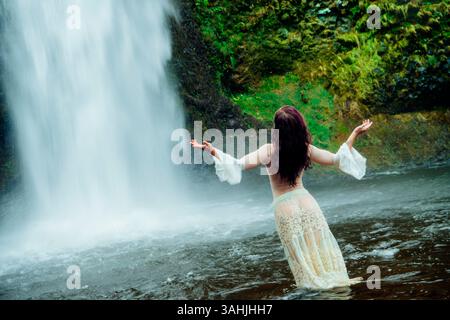 Donna in piedi in acqua vicino a una cascata, braccia sollevate, circondata da vegetazione lussureggiante. Silver Falls, Oregon, Stati Uniti Foto Stock