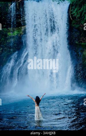 Donna in piedi in acqua con le braccia sollevate davanti a una grande cascata. Silver Falls, Oregon, Stati Uniti Foto Stock