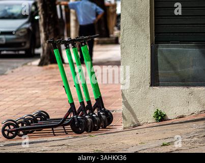 Una fila di scooter elettrici verdi luminosi parcheggiati ordinatamente su un marciapiede della città, con la vita urbana che si svolge sullo sfondo Foto Stock
