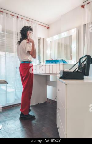 Una persona non binaria che indossa pantaloni rossi e una t-shirt bianca sta indossando il trucco in uno spogliatoio luminoso, mettendo in risalto temi di autoespressione, Foto Stock