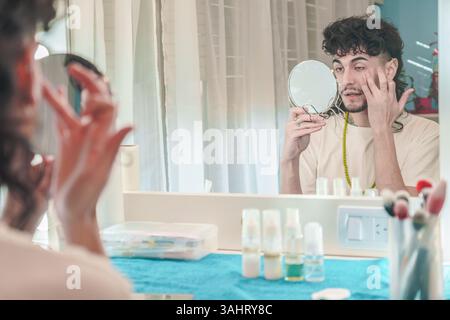 Il giovane con i capelli ricci si sta truccando davanti a uno specchio su un tavolo da toilette, tenendo un piccolo specchio rotondo in una mano e applicando il trucco wi-fi Foto Stock