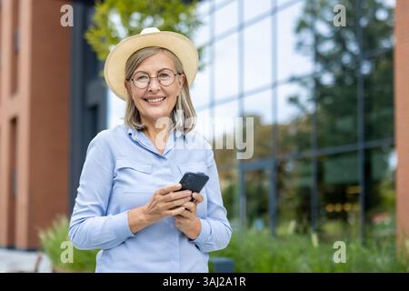 Un'elegante donna anziana con occhiali che indossano un cappello, sorride tenendo il telefono all'aperto in un ambiente moderno, mostrando soddisfazione. Foto Stock