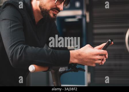Un giovane uomo urbano elegante con scooter elettrico in piedi di fronte al moderno edificio aziendale sulla strada della città e utilizzando un telefono. Foto Stock