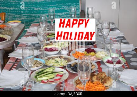 un tavolo con cibo e bicchieri davanti al quale è scritto "buone feste" e uno sfondo di una festa di compleanno con colori, blu pallido e rosso Foto Stock