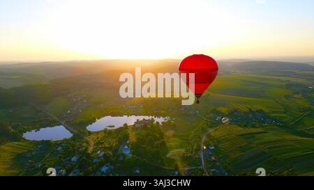 Mongolfiera sulla campagna al tramonto. Una vista aerea mozzafiato di una mongolfiera rossa che sorvola una lussureggiante campagna all'alba. Foto Stock