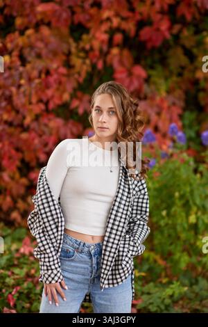 Autunno Ritratto alla moda: Giovane donna con un outfit casual che si posa contro la vivace parete di edera rossa in un ambiente urbano all'aperto. Eleganti vetrine in stile stagionale Foto Stock
