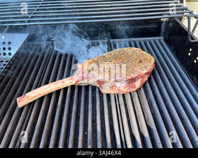 Bistecca di tomahawk condita sfrigolante su una griglia calda con lievitazione del fumo, che offre le classiche vibrazioni del barbecue all'aperto e preparazioni di carne di prima qualità. Foto Stock