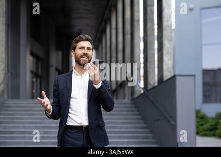 Un uomo d'affari ben vestito sorride mentre parla al telefono vicino a un edificio urbano. Le sue mani gestiscono come se stesse enfatizzando un punto. Foto Stock