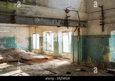 Un'area soleggiata coperta di sabbia in una storica fabbrica abbandonata, caratterizzata da grandi finestre ad arco. Ideale per la rappresentazione del degrado urbano e del patrimonio industriale. Foto Stock