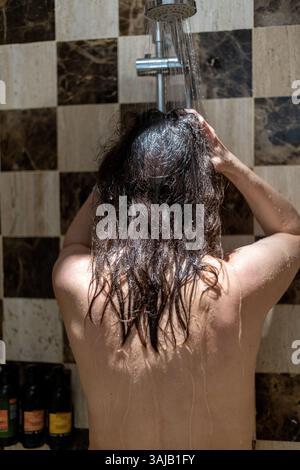 Una persona è nella doccia, lavandosi i capelli mentre l'acqua scorre dalla doccia a soffitto. Lo sfondo presenta una moderna parete piastrellata in tonalità tenui Foto Stock