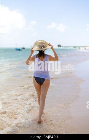 Una giovane donna cammina a piedi nudi sulla spiaggia sabbiosa, tenendo il cappello da sole, con dolci onde che le lambiscono ai piedi. Il cielo azzurro e limpido completa il seno Foto Stock