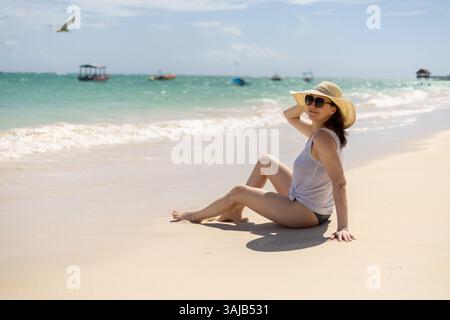 Una donna si rilassa su una spiaggia soleggiata, indossando un cappello di paglia e occhiali da sole. Si siede sulla sabbia calda, osservando le onde e le barche nelle vicinanze. Il cielo limpido Foto Stock