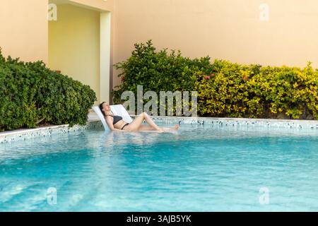 Una giovane donna gode di un momento tranquillo rilassandosi presso una piscina blu brillante, prendendo il sole in un lussureggiante giardino. Il verde vivace aggiunge al tran Foto Stock