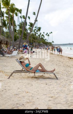 Punta Cana, Repubblica Dominicana - 25 marzo 2025: Una donna in bikini si rilassa su una sedia a sdraio sotto un cappello solare, mentre gli amanti della spiaggia passeggiano lungo la sabbia Foto Stock