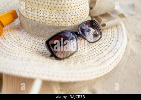 Un cappello di paglia a tegole larga adornato da un nastro si appoggia sulla sabbia calda insieme a eleganti occhiali da sole neri e crema solare, perfetto per una spiaggia soleggiata Foto Stock