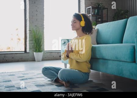 Una giovane donna che meditava a casa indossando le cuffie e un casual top giallo in un soggiorno splendidamente illuminato con un'atmosfera rilassante. Foto Stock