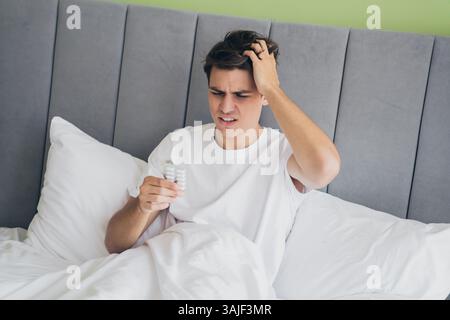 Un giovane che si sveglia a letto tenendo le medicine, sembra confuso e si gratta la testa in una luminosa camera da letto mattutina. Foto Stock
