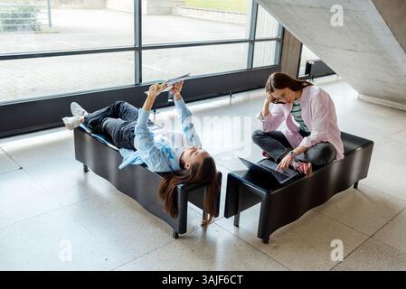 Studentesse che studiano insieme nel corridoio dell'università, sedute e sdraiate su due panche di legno Foto Stock