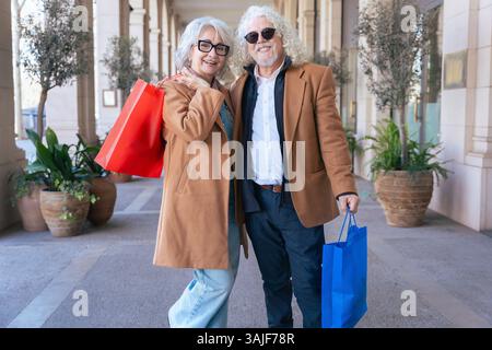 La coppia senior sorride alla macchina fotografica mentre abbraccia e tiene in mano borse colorate Foto Stock