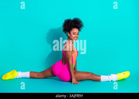 Giovane donna atletica che sorride con sicurezza indossando un vivace abbigliamento fitness mentre si mette in posa su uno sfondo turchese brillante Foto Stock