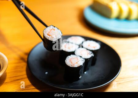 Un primo piano di sushi su un piatto nero, con le bacchette che raccolgono un rotolo. Il sushi presenta ripieno di riso e gamberi, con uno sfondo di addi Foto Stock
