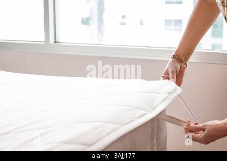 mani di donna che preparano il letto, si infilano in coprimaterasso e lenzuola in una camera illuminata con grande finestra. Pulizia delle camere dell'hotel. Foto Stock