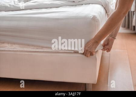 mani di donna che preparano il letto, lenzuola e biancheria da letto in una camera illuminata con grande finestra. Pulizia delle camere dell'hotel. Foto Stock