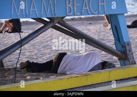 Persona che dorme sulla spiaggia della Florida, circondata da sabbia e mare, luce calda, povertà urbana, lotta umana, stile documentario di viaggio Foto Stock