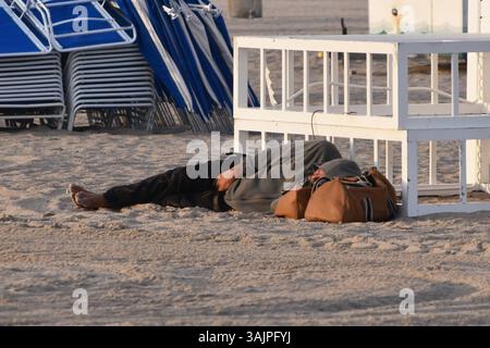 Dormiente sulla spiaggia sabbiosa di Miami, mare alle sue spalle, calda atmosfera costiera, problemi sociali, realtà della vita in spiaggia, una scena di stile street Foto Stock