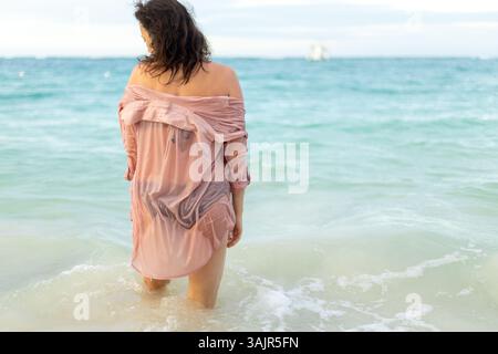Una donna cammina a piedi nudi tra le onde poco profonde di una spiaggia, godendosi l'acqua fresca. Indossa una copertura fluida e guarda attentamente all'orizzonte come Foto Stock