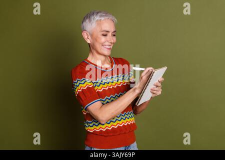 Donna anziana che scrive su un notebook su sfondo verde mentre sorride e si gode un momento casual Foto Stock