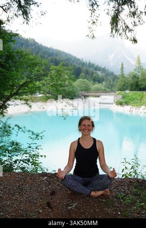 Donna che meditava tranquillamente all'aperto in un ambiente naturale tranquillo accanto a un tranquillo lago turchese, circondato da vegetazione lussureggiante e montagne. Si siede Foto Stock