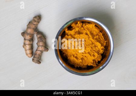 Vista dall'alto della pasta di curcuma grezza in una ciotola di acciaio su una superficie di legno chiaro, con due radici di curcuma grezze intere poste accanto ad essa. Foto Stock
