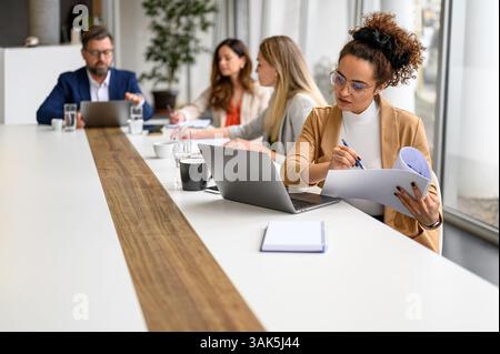 Donna d'affari sicura che esamina documenti e lavora su un notebook mentre i colleghi collaborano in sala riunioni Foto Stock