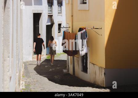 Lisbona, Portogallo - 12 luglio 2024: Una giovane coppia passeggia attraverso uno stretto vicolo acciottolato a Lisbona, Portogallo, dove il bucato è appeso su un clothesli Foto Stock