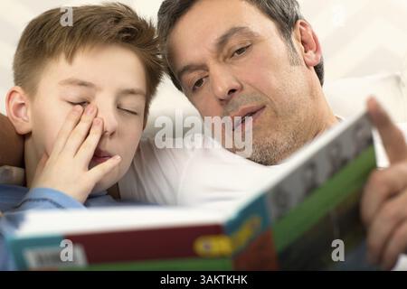 Inquadratura ravvicinata di un padre la lettura di un libro per il figlio che è già addormentata e strofinando i suoi occhi Foto Stock