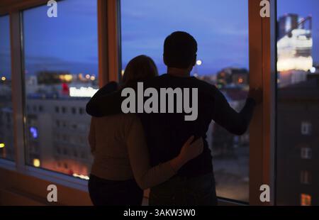 Torna shot di giovani amare giovane dalla finestra la notte. Esse abbracciando e godendo la vista della citta'. Momenti romantici Foto Stock