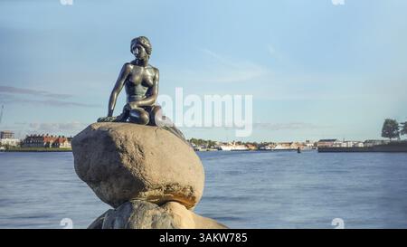 Vista frontale della statua della Sirenetta su grandi massi in Danimarca con il porto sotto il cielo blu sullo sfondo Foto Stock