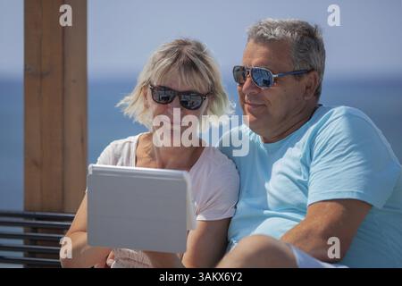 Una coppia di mezza età in occhiali da sole guarda il loro ipad, seduto su una panchina con una vista sul mare Foto Stock