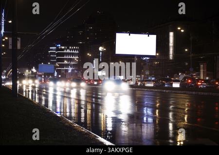 Illuminato bianco vuoto billboard sul lato di una strada con le auto in movimento e un blocco di appartamenti in background, di notte Foto Stock
