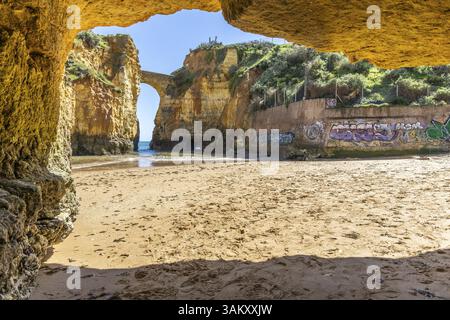 Rocce gialle e scogliere su una spiaggia sabbiosa. Una baia sul mare, sopra la quale il vecchio ponte in pietra della fortezza di Pinhao collega due scogliere. Praia da Cald Foto Stock