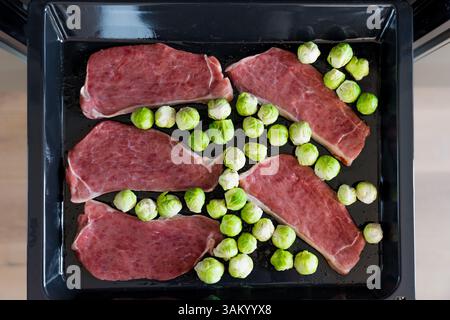 Bistecche di maiale crude e cavolini di Bruxelles su vassoio da forno pronti per la tostatura in cucina. Messa a fuoco selettiva Foto Stock